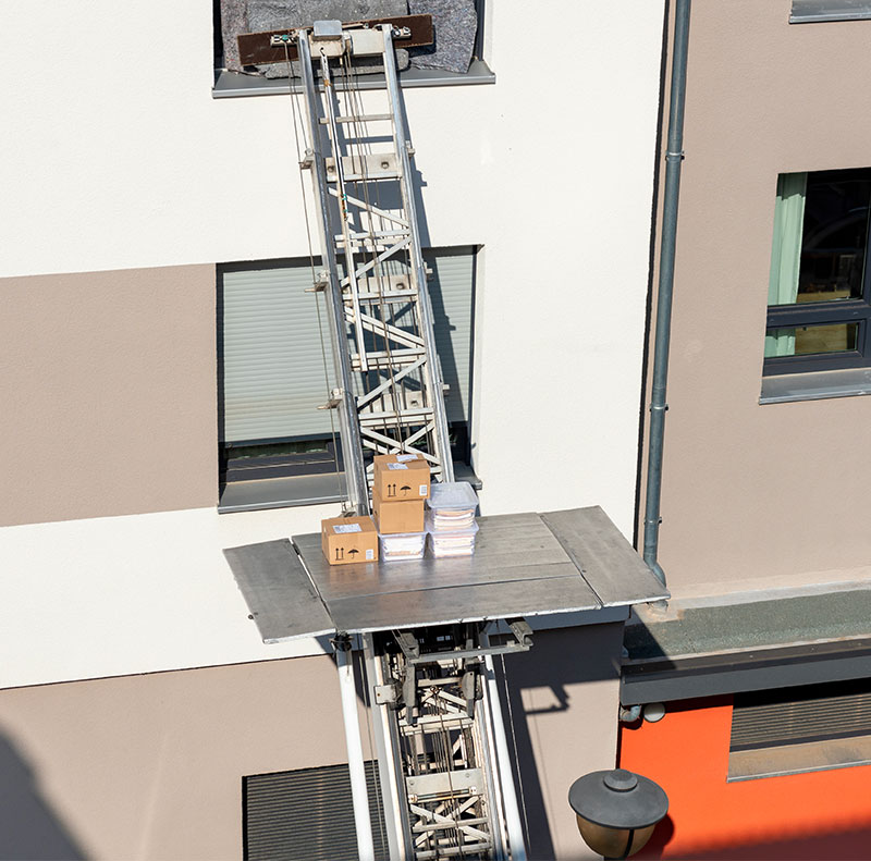 La piattaforma di un'autoscala in funzione sale lungo la facciata di un palazzo moderno, trasportando scatoloni e oggetti imballati per il trasloco