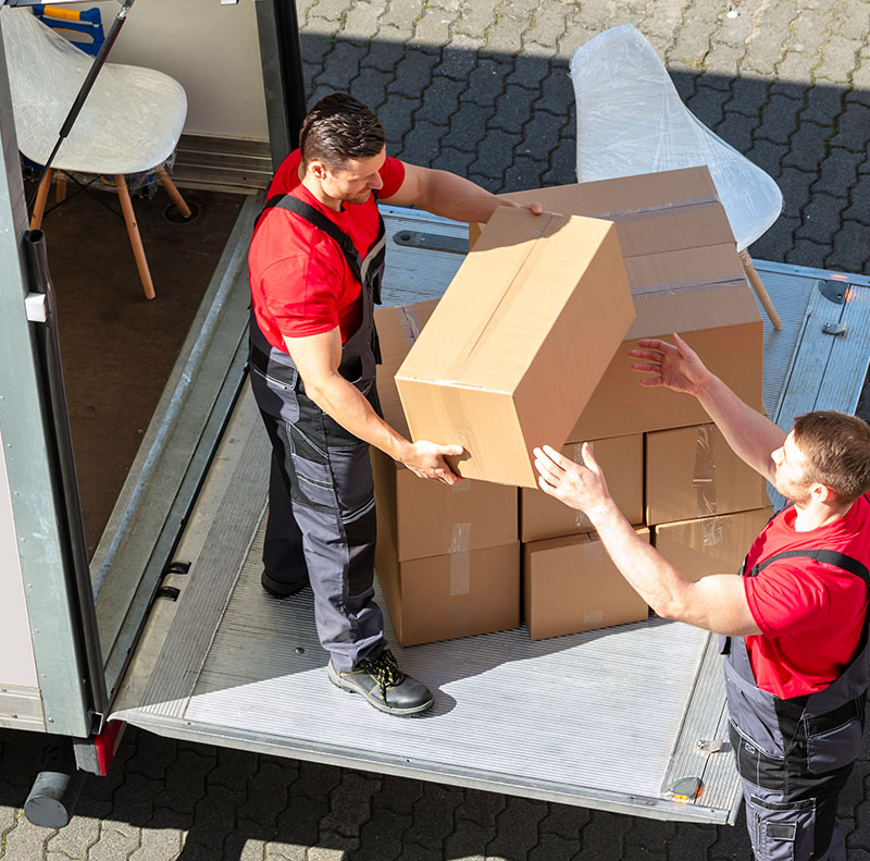 Vista dall'alto su due facchini in divisa che lavorano in coordinamento sulla pedana del furgone a noleggio per sollevare e caricare uno scatolone di cartone