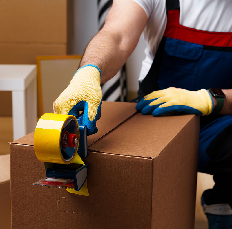 Primo piano sulle mani guantate di un addetto che utilizza un dispenser professionale per sigillare con cura una scatola di cartone con nastro adesivo