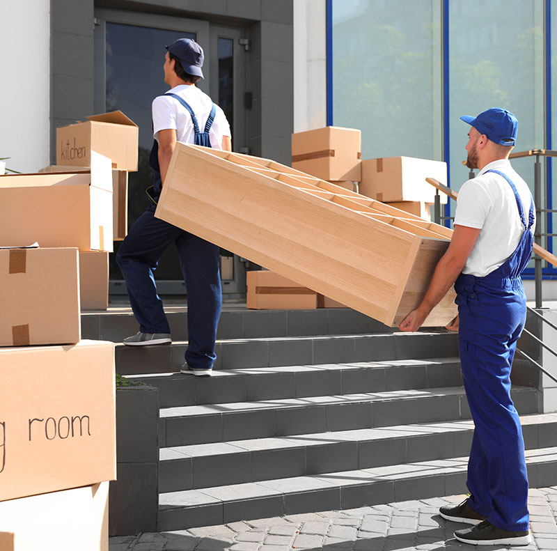 Due addetti ai traslochi trasportano manualmente un mobile in legno su una scalinata esterna circondati da scatoloni etichettati per il trasferimento